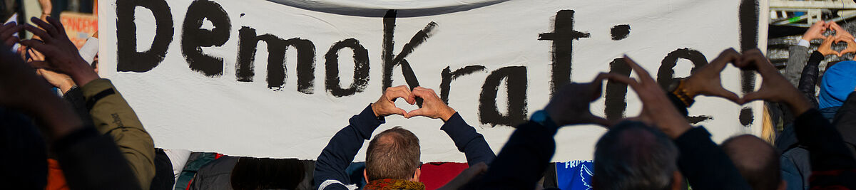 Foto mit Menschen, die ihre Hände in die Höhe strecken und ein Banner mit dem Schriftzug Demokratie halten