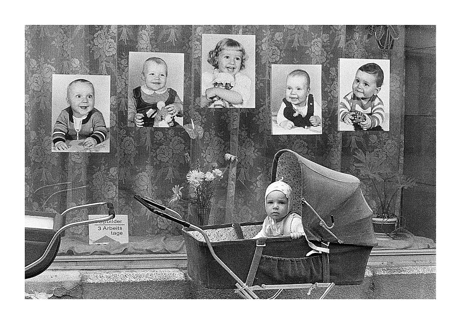 ein Kleinkind sitz in einem Kinderwagen vor einem Schaufenster mit Fotografien von Kinderporträts