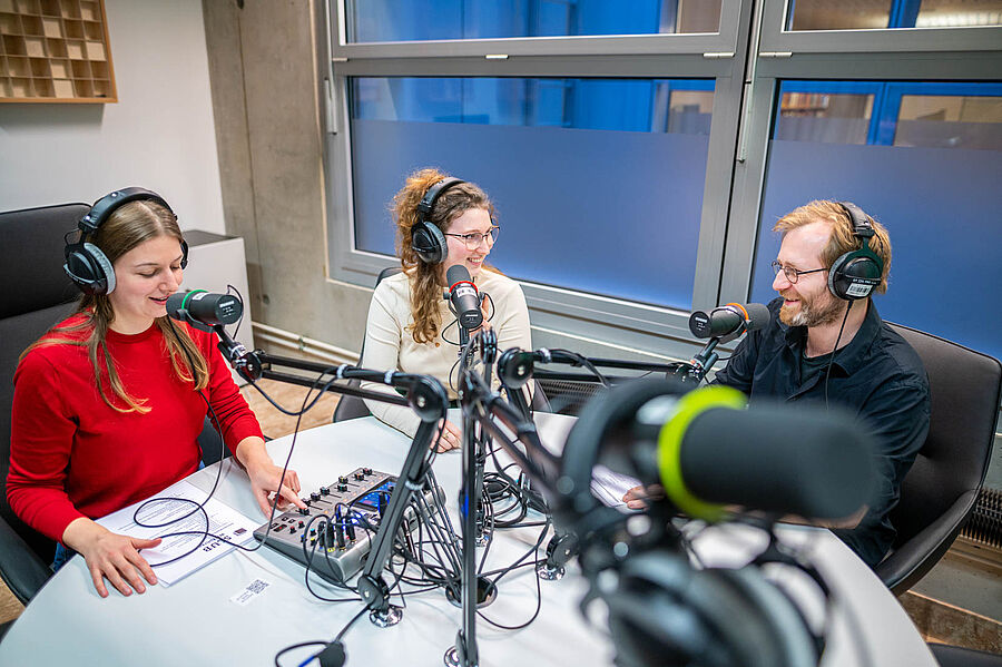 Drei Menschen sitzen im Podcaststudio und reden. Runder Tisch mit Technik und sechs Mikrofonen. 